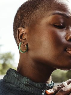 Close-up of a woman with short hair wearing a green top and showcasing a pair of gold hoop earrings with green beaded accents. Her face is partially visible with her eyes gently closed, exuding a calm and confident expression. Her hand, adorned with a white stone ring, rests softly near her neck, adding an elegant touch to the composition. The background is softly blurred, keeping the focus on the jewelry and her serene expression. Bohemian Large Beaded Dangle Hoop Earrings, Modern Wire Wrapped Hoop Earrings, Chic Green Hoop Jewelry, Luxury Handmade Green Hoop Earrings, Unique Green Hoop Earrings, Bohemian Large Beads Multicolor Hoop Earrings, Modern Turquoise Hoop Earrings, Best Travel Accessories, Luxury Gold-tone Brass Hoop Earrings