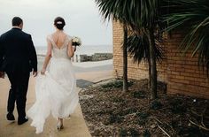 the bride and groom are walking down the sidewalk towards the beach in their wedding attire