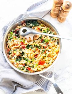 a skillet filled with pasta and vegetables on top of a white tablecloth next to wooden spoons