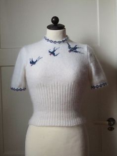 a white sweater with blue birds on the collar is displayed in front of a door