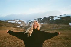 a woman standing in the middle of a field with her arms spread out and snow covered mountains behind her