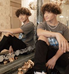 two young men sitting in front of a mirror with gold jewelry on the floor next to them