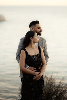 a man and woman standing next to each other in front of the ocean with their arms around each other