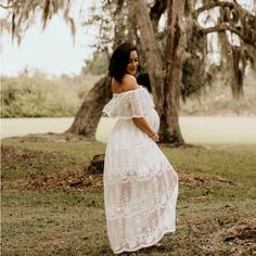 a pregnant woman standing in front of a tree wearing a white lace dress with off the shoulder sleeves