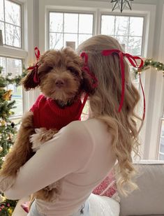 a woman holding a small dog in her arms near a christmas tree and window sill