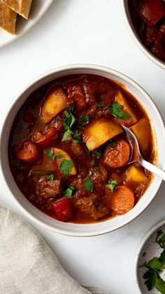 two bowls filled with stew and vegetables on top of a white tablecloth next to bread