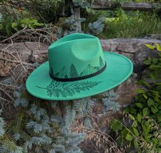 This hand burnt wide brim fedora makes a perfect addition to any nature lover's  wardrobe. The Kelly green material burns a gorgeous shade of emerald green allowing the design to stand out. I burnt pine trees in front of a mountain scene around the entire crown of the hat. Each mountain is intricately burned by hand with a pyrography machine using various tips to achieve depth and a rock-like texture.  On the right side brim of the hat I burnt a realistic fern. For a finishing touch, I burnt dis Custom Handmade Hats For Outdoors, Handmade Green Bohemian Fedora, Handmade Curved Brim Hat For Outdoors, Handmade Curved Brim Hat For Outdoor, Handmade Flat Brim Hats For Outdoor, Handmade Green Fedora With Curved Brim, Handmade Green Fedora With Short Brim, Green Handmade Fedora With Short Brim, Custom Brimmed Fedora For Outdoor