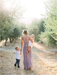 a mother and her two children walking through an olive grove in the sun with their arms around each other