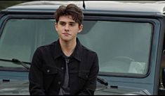 a young man sitting on the hood of a car in front of a parked truck