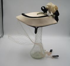 This is a vintage 1950s, fascinator, ladies hat.  The hat is adorned with a beautiful white rose in its posterior section.  The hat is highlighted by a black velvet ribbon, and it is trimmed by a black velvet piping which circles around the top of the hat.  This hat is further enhanced by a nylon face veil which ties behind the head.  This hat has firm clips/anchors on each side to help cradle the hat on one's head.  Beautiful cream/ivory color.  Fully intact.  Nice and clean for its age.  The inner circumference of this hat is approximately 22 inches.  The hat measures 6 1/2 inches wide and 8 1/2 inches in length.  This hat was purchased by my mother in the 1950s after she came to the U.S.A. from Poland. 1950s Fascinator, Vintage White Hat With High Crown, Victorian Hat With Veil, Vintage Cream Hat For Vintage Events, White Vintage Cloche Fascinator, Vintage White Hat Headpiece, Princess Story, Flower Piping, 1950s Woman