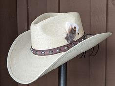 Chet starts with a Mexican soft palm leaf hat body that has a 3 3/4" brim and 5 1/4" crown. The band is simple with distressed brown leather and antique nickel color spots. The back of the band has a deer antler bead for accent. A neutral feather combo completes the hat. Note: Please allow 3-4 weeks. Fitted Rustic Hat For Country Events, Rustic Fitted Fedora For Country Events, Natural Color Adjustable Western Hat Bands, Adjustable Natural Color Western Hat Band, Adjustable Western Hat Band In Natural Color, Distressed Brown Adjustable Hat For Western-themed Events, Adjustable Natural Western Hat Bands, Rustic Natural Hats For Western-themed Events, Rustic Natural Hat For Western-themed Events