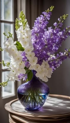 purple and white flowers in a blue glass vase on a table next to a window