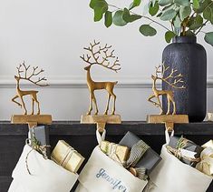 three christmas stockings with reindeer decorations on them and presents wrapped in gold foil sitting next to a black vase filled with greenery