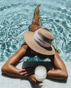 a woman in a straw hat sitting on the edge of a swimming pool next to a drink