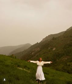 a woman standing on top of a lush green hillside under a cloudy sky with her arms outstretched