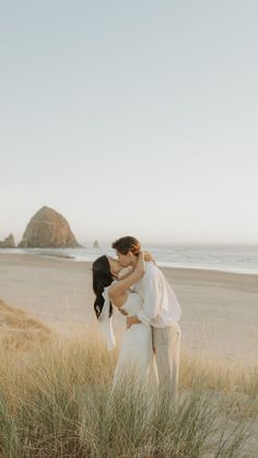 a man and woman kissing on the beach in front of some tall grass with an ocean in the background