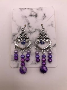 2 Inch drop-earrings made with hypoallergenic materials.   Beads are attached to a silver plated heart charm consisting of a purple marble bead and three smaller glass beads in three different shades of purple/lavender in the center.  Left/Right sides contain the same smaller glass beads in the three different shades of purple/lavender as the center.  The middle of the heart charm is accented with four tiny glass beads as an accent.  These beads are a darker shade of purple. Includes soft ear backings. Materials are environmental, lead free and non-toxic. Light weight, smooth surface, and a touch of sheen to add elegance. The earrings are attached to a marble cardboard earring holder and are shipped in a sealed plastic cover, and placed inside a mesh drawstring "thank you" pouch as shown. Nickel-free Purple Dangle Chandelier Earrings, Purple Heart-shaped Beaded Jewelry, Purple Metal Dangle Earrings, Purple Dangle Metal Earrings, Purple Dangle Heart Earrings For Pierced Ears, Nickel-free Purple Chandelier Earrings Gift, Purple Drop Chandelier Earrings As Gift, Dangle Heart Bead Earrings, Metal Dangle Heart Earrings With Heart Beads