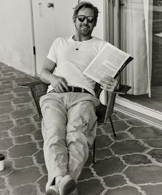 a man sitting in a chair holding a book and smiling at the camera while wearing sunglasses