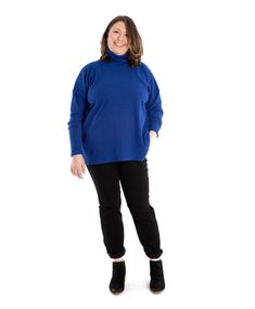 a woman standing in front of a white background wearing a blue sweater and black pants