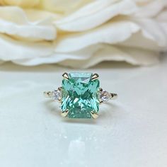 "The ring pictured is lab created green paraiba with moissanite accents #7156 Rose Gold is available upon request as a custom order and is a final sale (non refunable, non returnable, non exchangeable). -Approximate total carat weight: approx. 5.40ctw diamond equivalent -Center Stone Size: 11x9mm - approx. 5.00ct diamond equivalent -Center Stone Shape: radiant -Gem Type: lab created paraiba -Stone Clarity: VS2 -Stone Color: Light/Mint Green -Moh's Scale: 7.5-8 hardness -Side Stones Size: Pear 5x Green Moissanite Jewelry In Radiant Cut, Green Moissanite Jewelry With Radiant Cut, Green Moissanite Radiant Cut Jewelry, Green Radiant Cut Moissanite Jewelry, Green Aquamarine Jewelry With Center Stone, Gia Certified Aquamarine Wedding Jewelry, Gia Certified Aquamarine Jewelry For Wedding, Green Moissanite Jewelry For Promise, Three Stone Wedding Ring