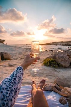 a person holding a glass of wine while sitting on a towel near the ocean at sunset