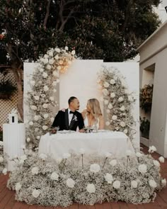 a man and woman sitting at a table in front of a white backdrop with flowers