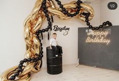 a young boy sitting on top of a black barrel in front of balloons that spell happy birthday