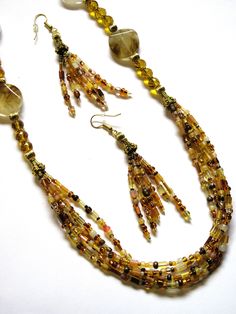 Shades of brown, beige and gold seed beads were used to make this one-of-a-kind jewelry set. The bottom of this 26-inch necklace is a grouping of 5 strands of mixed seed beads with antique gold bead caps at each end. The sides are gold faceted glass rondelles mixed with gold plated corrugated rounds, gold plated spacers, and tiger skin glass. The gold-plated chain could be used to shorten the necklace by fastening the clasp higher up the chain. The seed bead tassel earrings dangle about 2.5 inches from the loops of the gold-plated wires. This jewelry set will be slipped into an organza bag and shipped in a bubble mailer. Bohemian Gold Earrings With Polished Beads, Gold Multi-strand Gemstone Beads, Multi-strand Brown Beads For Gifts, Brown Multi-strand Beads For Gift, Artisan Gold Beaded Necklaces With Dangling Beads, Unique Gold Beaded Necklace With Dangling Beads, Brown Multi-strand Beaded Necklace With Gold Beads, Brown Gold Beads Multi-strand Necklace, Gold Multi-strand Dangling Beads