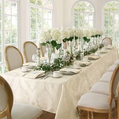 a long table with white flowers in vases and plates on it is surrounded by windows