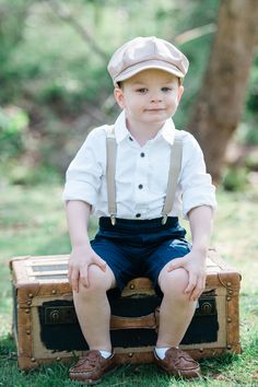 a young boy sitting on top of an old trunk in the grass wearing suspenders and a hat