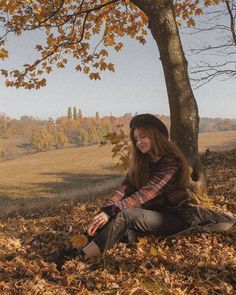 a young woman sitting under a tree in the fall with leaves on the ground around her