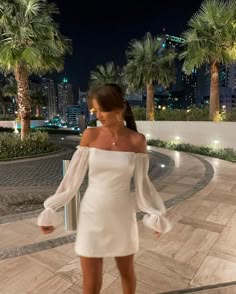 a woman in a white dress is standing on the sidewalk with palm trees behind her