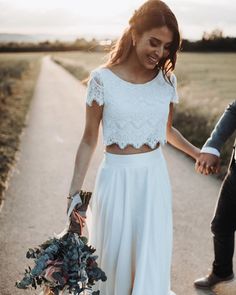 a man and woman holding hands walking down a road with flowers in their hand while the sun is setting