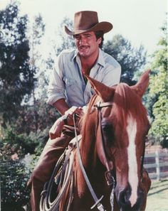 a man riding on the back of a brown horse next to a white fence and trees