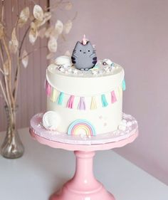 a white cake with a cat on top sitting on a pink stand next to a vase