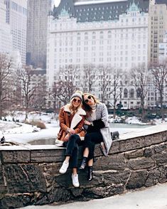 two women are sitting on a stone wall in the snow, with large buildings behind them