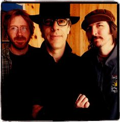 three men standing next to each other in front of a wooden wall and wearing glasses