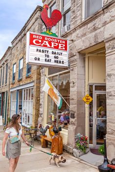 a woman walking her dog down the street in front of a cafe with a sign that says red rooster cafe