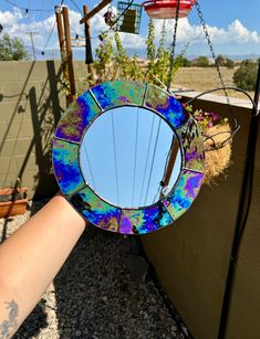 a hand holding up a colorful mirror hanging from the side of a building next to a bird feeder