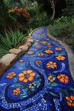 a garden path made out of blue and orange tiles with flowers on the side, surrounded by greenery