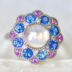 Rainbow Moonstone Sapphire Halo Gold Ring Pink and Blue Sapphire Edwardian Style moonstone Ring wedding Engagement Ring. Rainbow Moonstone Sapphire Halo Ring Pink and Blue Sapphire  Edwardian Style moonstone Ring wedding Engagement Ring. 14K White Gold US7 This cocktail ring features my mini cocktail ring designed with one piece of rainbow color  moonstone cabochon and a fancy double halo of cornflower blue sapphire and pink sapphires! The size is in US 7 made of solid 14K white gold can be size Blue Celestial Moonstone Ring, Celestial Blue Round Moonstone Ring, Blue Moonstone Ring With Accent Stones, Celestial Blue Moonstone Ring, Sapphire Moonstone Gemstone Ring, White Multi-stone Moonstone Ring, Blue Moonstone Ring With Accent Stones For Anniversary, Blue Moonstone Promise Ring, Unique Moonstone Ring With Accent Stones