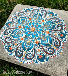 a blue and orange painted stone with an intricate flower design on the top, sitting in grass