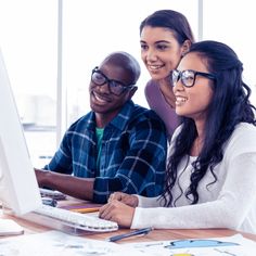 three people sitting at a desk looking at a computer screen and smiling for the camera