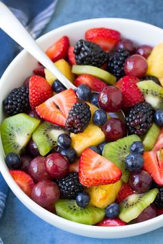 a white bowl filled with fresh fruit on top of a blue table cloth next to a spoon