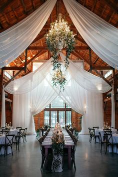 the inside of a barn with tables and chandeliers hanging from the ceiling, surrounded by white drapes