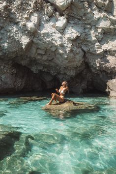 a woman sitting on top of a rock in the ocean next to a rocky cliff