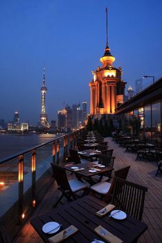 an outdoor dining area with tables and chairs overlooking the water at night, in front of a city skyline