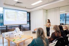 a group of people sitting around a table in front of a projector screen with a woman giving a presentation