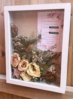 a white frame with some flowers in it on a wooden wall next to a piece of paper