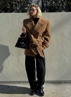 a woman standing next to a white wall holding a black purse and wearing a brown coat